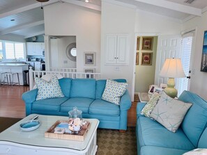 A living room with a blue sofa set, a coffee table with decorative items, and a kitchen area in the background. - Carpe Diem 4 Bedroom Home by RedAwning (Supply)