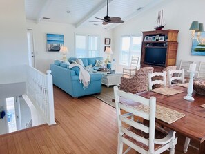 A spacious living room with a blue sofa, a dining table with chairs, a television, and a ceiling fan. - Carpe Diem 4 Bedroom Home by RedAwning (Supply)