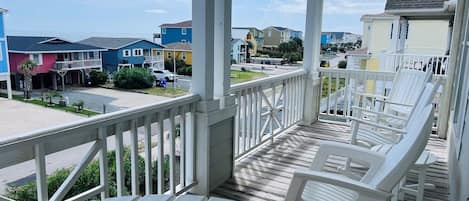 A covered porch with white chairs, a ceiling fan, and a view of colorful houses in the background.