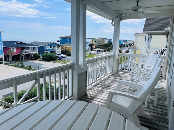 A covered porch with white chairs, a ceiling fan, and a view of colorful houses in the background.