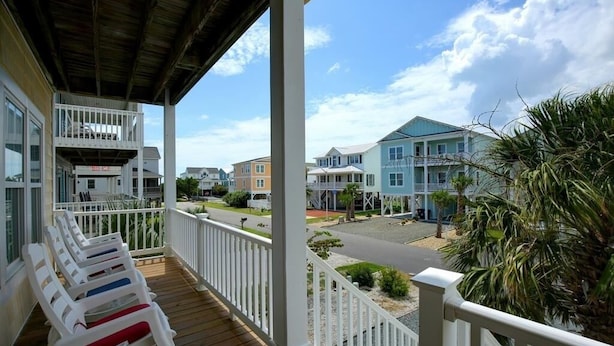 Um deck de madeira com guarda-corpos brancos e cadeiras de lounge, com vista para uma rua residencial com casas coloridas e um céu claro.