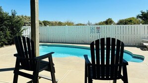 A backyard with a swimming pool, two Adirondack chairs, and a white fence.