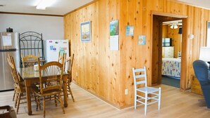 A wooden-paneled room with a dining table and chairs, a white chair, and a bed with a patterned bedspread. - Do-little Dock 3 Bedroom Home by RedAwning (Supply)