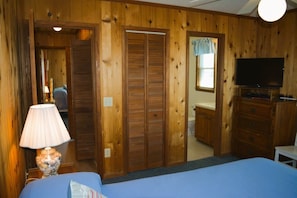 Una habitación con paneles de madera, una cama con ropa de cama azul, una lámpara, un televisor y un baño visible a través de una puerta abierta.