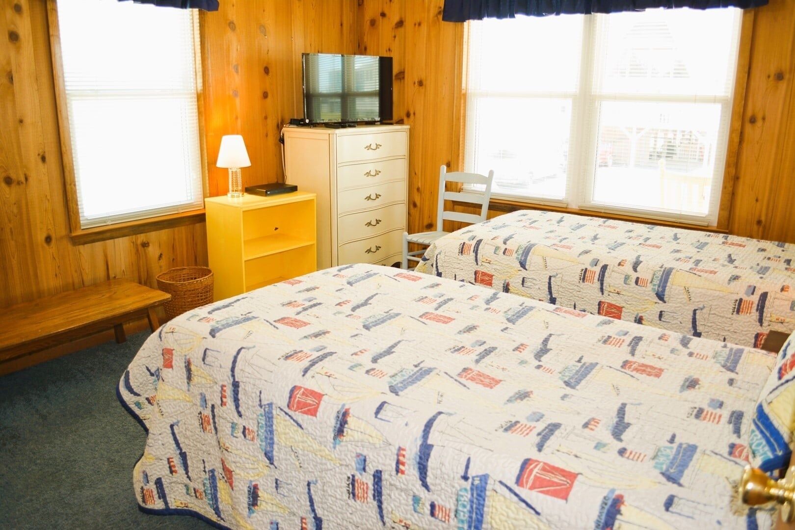 Ein Zimmer mit zwei Betten, einem Fernseher auf einer Kommode, einer Lampe und einem Fenster mit Jalousien.