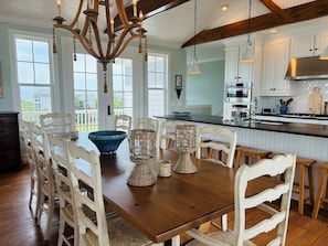 A dining area with a wooden table and white chairs, a chandelier, and a view of the outdoors through large windows. - Gray Goose 5 Bedroom Home by RedAwning (Supply)