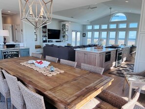A spacious kitchen with a large island, a dining area with a wooden table, and a view of the ocean through large windows. - Dune Escape 5 Bedroom Home by RedAwning (Supply)
