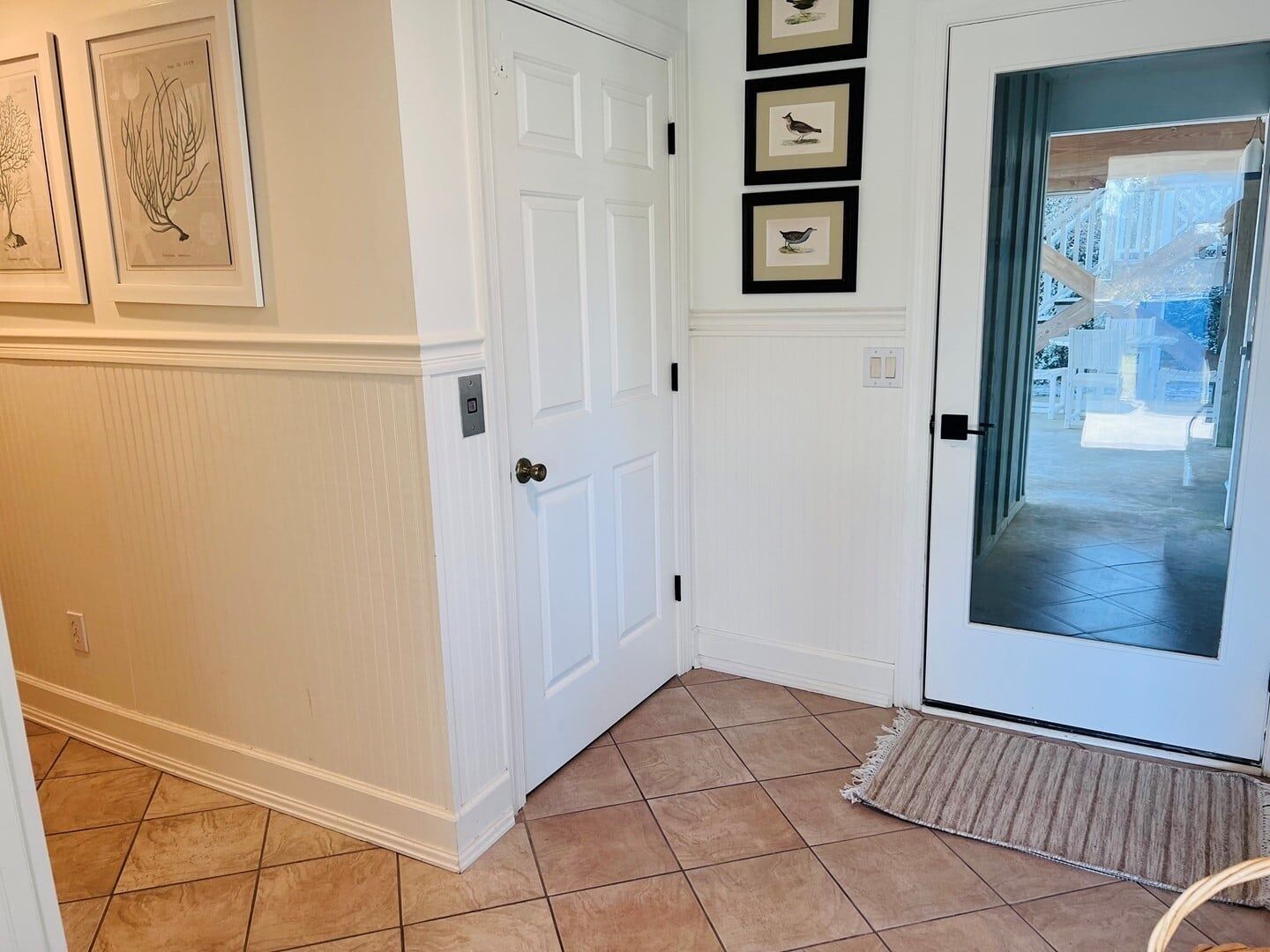 A hallway with a white door, framed botanical prints, and a tiled floor.