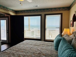 A bedroom with a large bed, two windows with blinds, a ceiling fan, and a view of a cityscape.