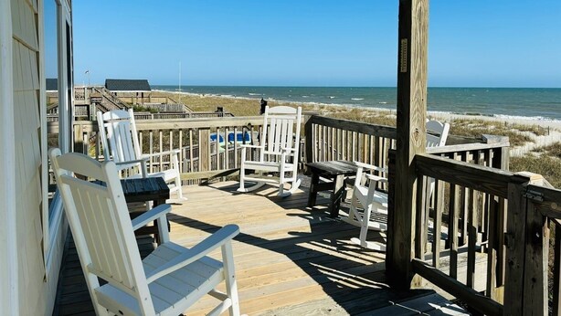 A wooden deck with white chairs and a table, overlooking a beach with a wooden boardwalk and beach huts in the distance. - Great Place 5 Bedroom Home by RedAwning (Supply)