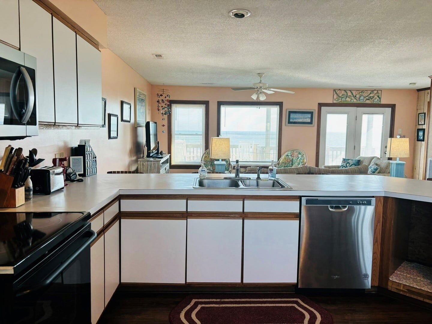 A modern kitchen with a dishwasher, microwave, and stove, a dining area with a table and chairs, and a view of the ocean through large windows.