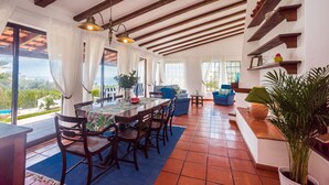 Un amplio salón con una mesa de comedor, sillas y un sofá. Grandes ventanales ofrecen vistas al exterior.