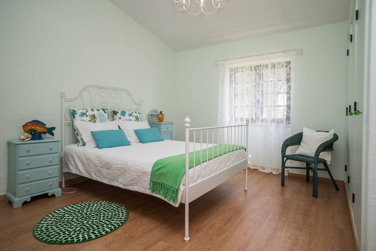 Un dormitorio ordenado con cama, cómoda, silla y ventana con cortinas translúcidas.