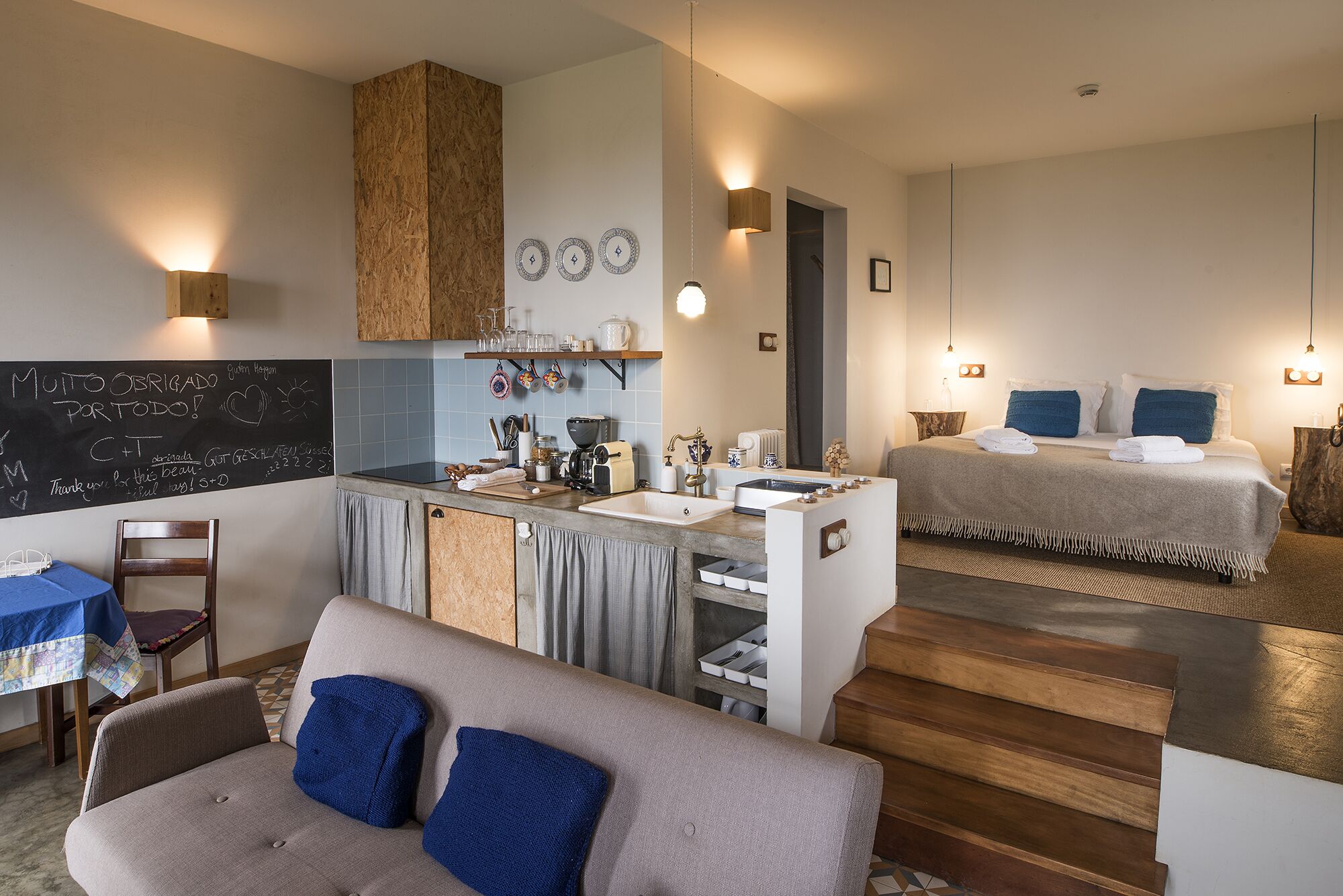 A compact hotel room with a kitchenette, a bed with blue pillows, and a dining area with a table and chair.