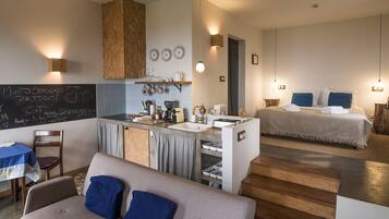 Una habitación de hotel compacta con kitchenette, una cama con almohadas azules y una zona de comedor con mesa y silla.