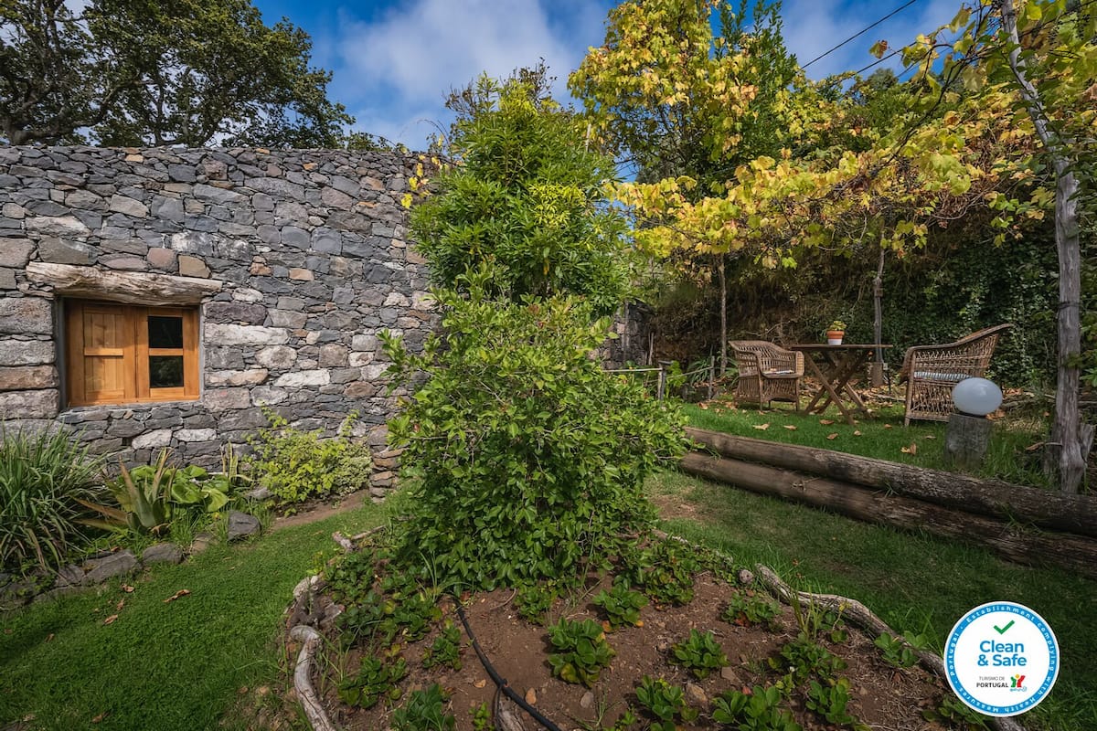 Un bâtiment en pierre avec une fenêtre en bois, entouré d’un jardin où poussent différentes plantes et agrémenté d’une petite table avec des chaises.