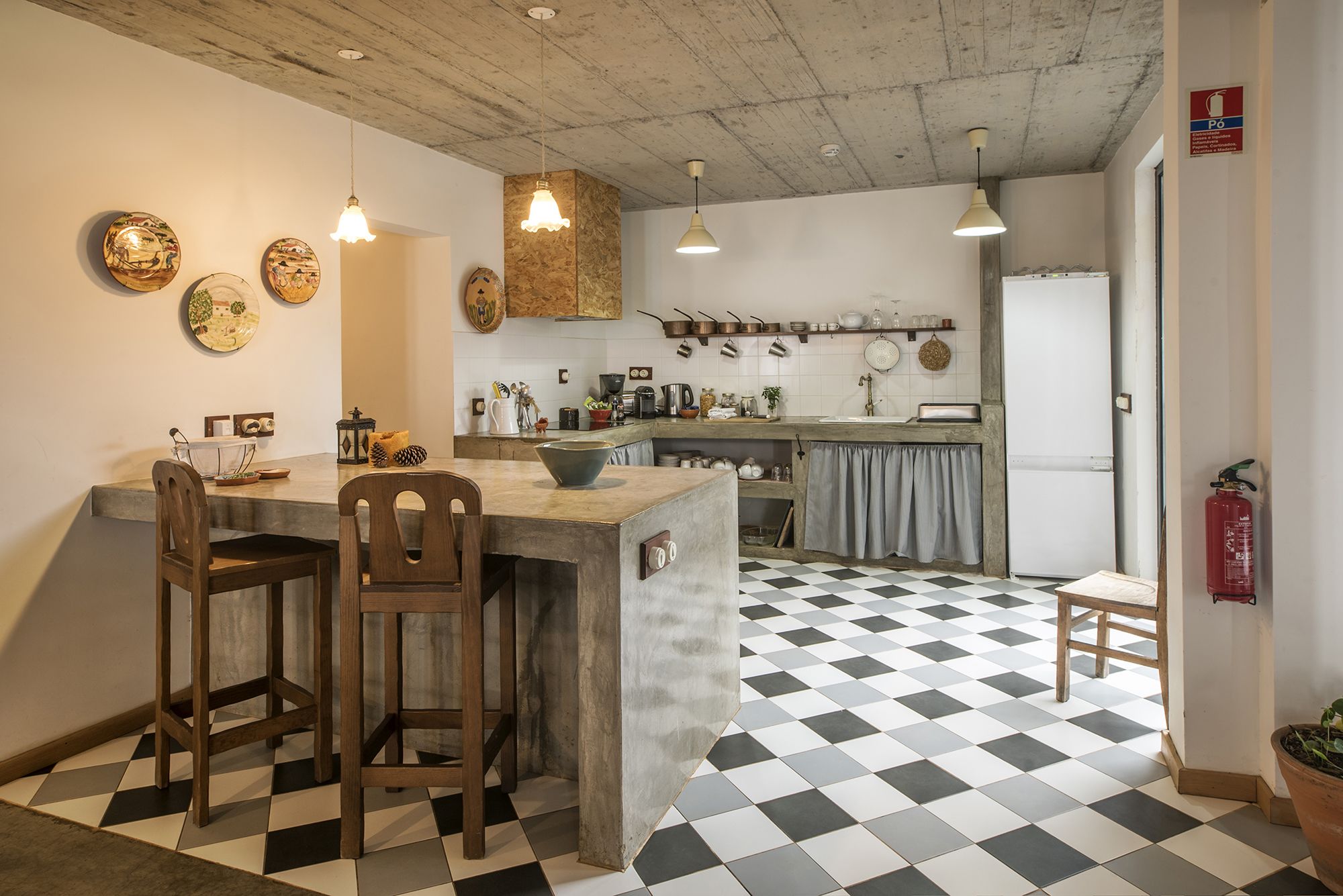 Een keuken met een geruit vloertje, houten stoelen, een bargedeelte, hangende lampen en diverse keukengerei.