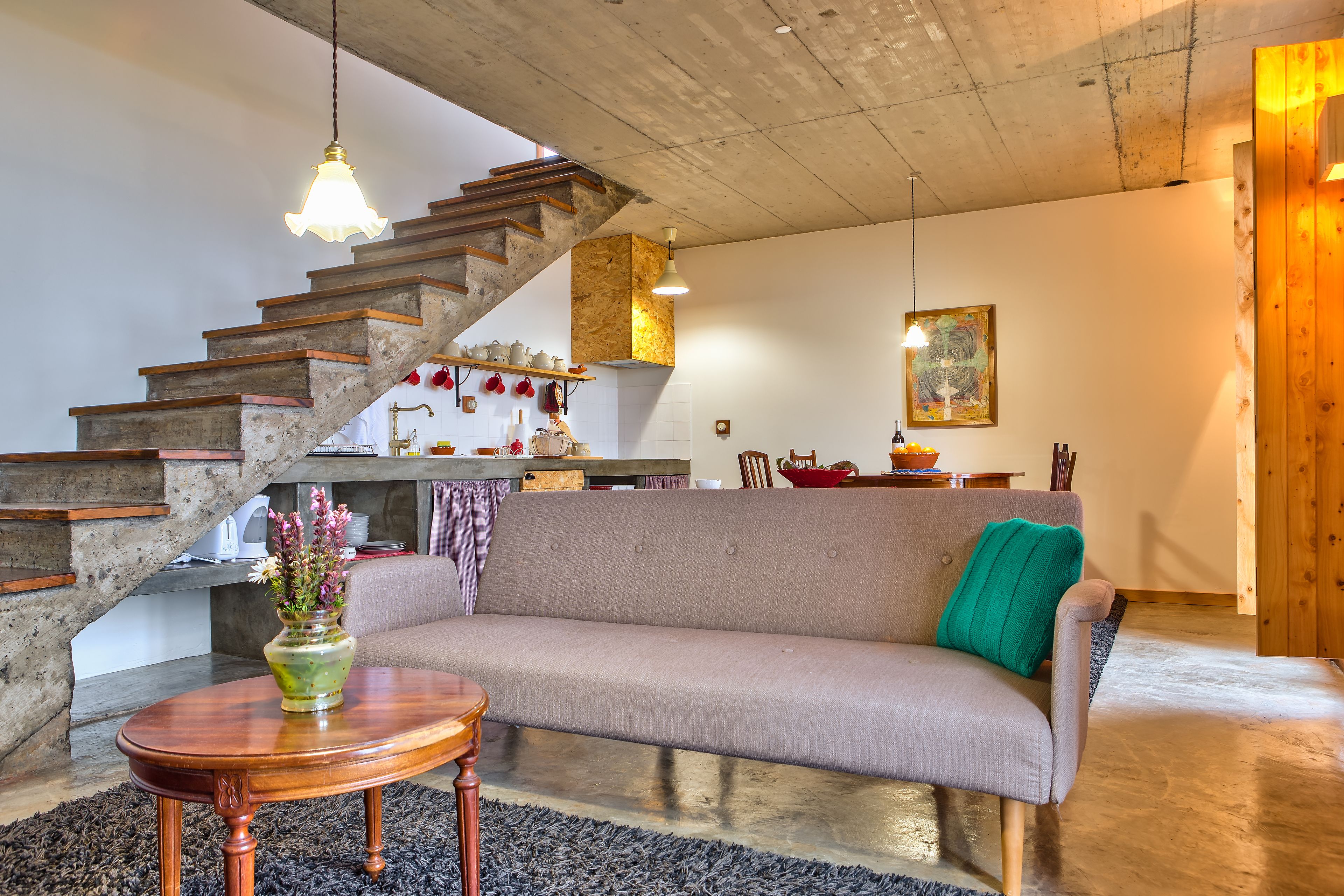 Una sala de estar con sofá, una mesa de madera, una escalera y una zona de cocina al fondo.