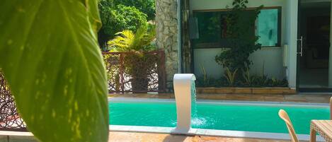 A swimming pool with a stone-tiled deck, a white column, and a view of a modern building with a window and a door.