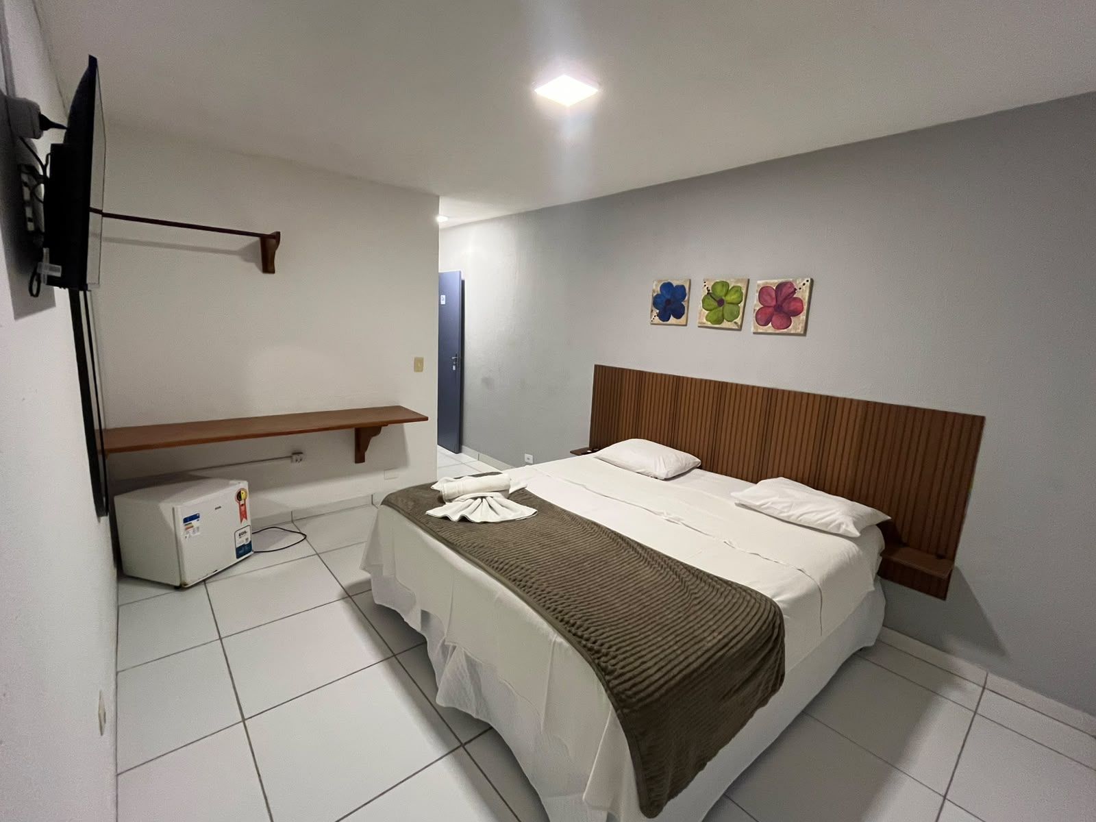 A hotel room with a bed, a TV, a shelf, and a wall with flower paintings.
