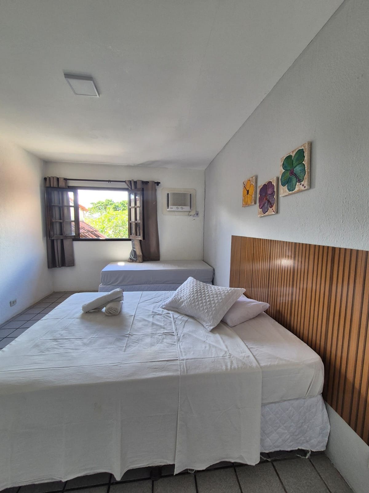 A bedroom with a bed, a window with curtains, an air conditioning unit, and a wall with three flower paintings.