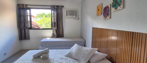 A bedroom with a bed, a window with curtains, an air conditioning unit, and a wall with three flower paintings.