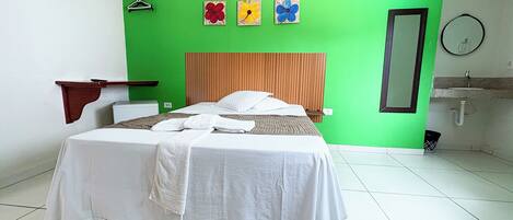 Una habitación de hotel con cama, cabecera de madera, pared de acento verde, espejo montado en la pared, mesita de noche y un perchero para toallas.