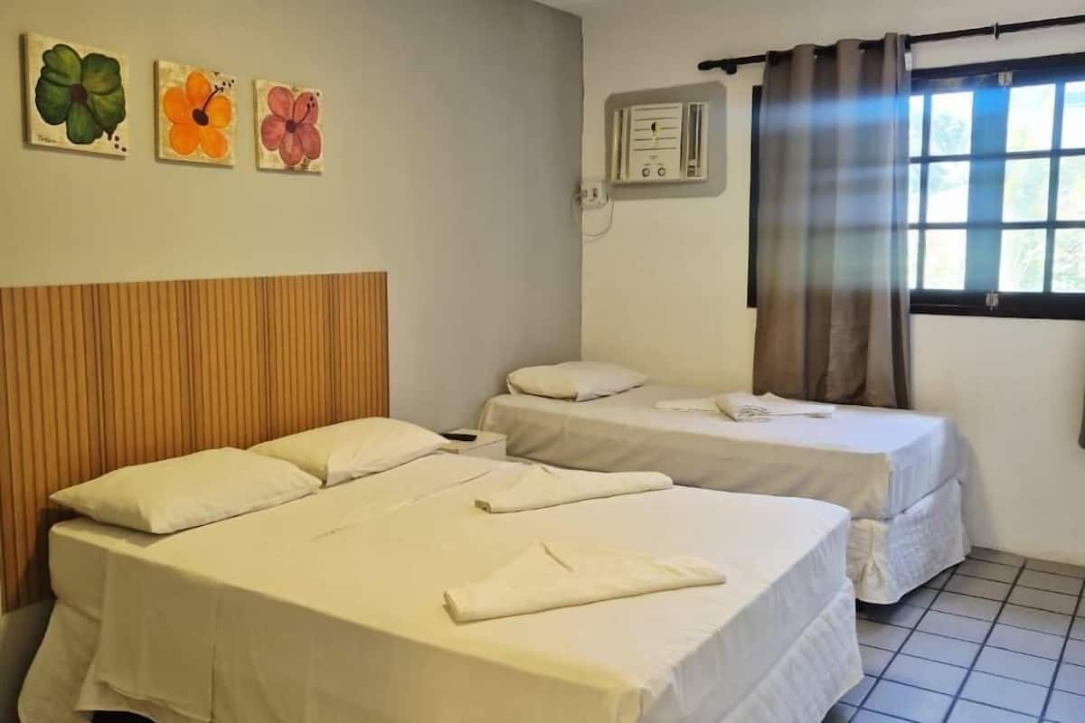 Quarto de hotel com duas camas, uma cabeceira de madeira, uma janela com cortinas e três quadros de flores na parede.