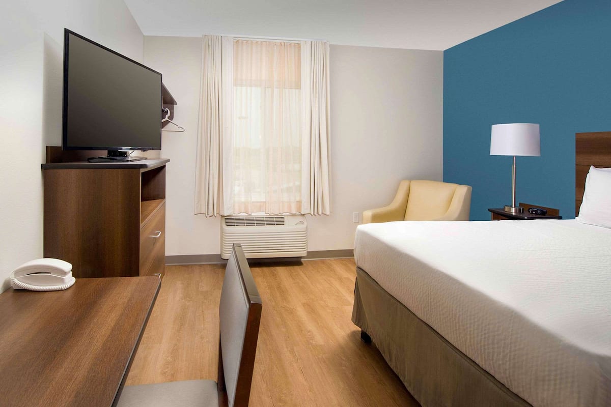Ett hotellrum med en säng, en tv, ett skrivbord med en telefon, en stol, en lampa och ett fönster med gardiner.