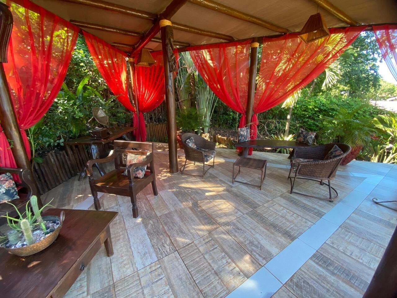 Un patio exterior con mobiliario de madera, cortinas rojas y una planta en maceta.