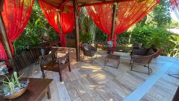 Un patio exterior con mobiliario de madera, cortinas rojas y una planta en maceta.