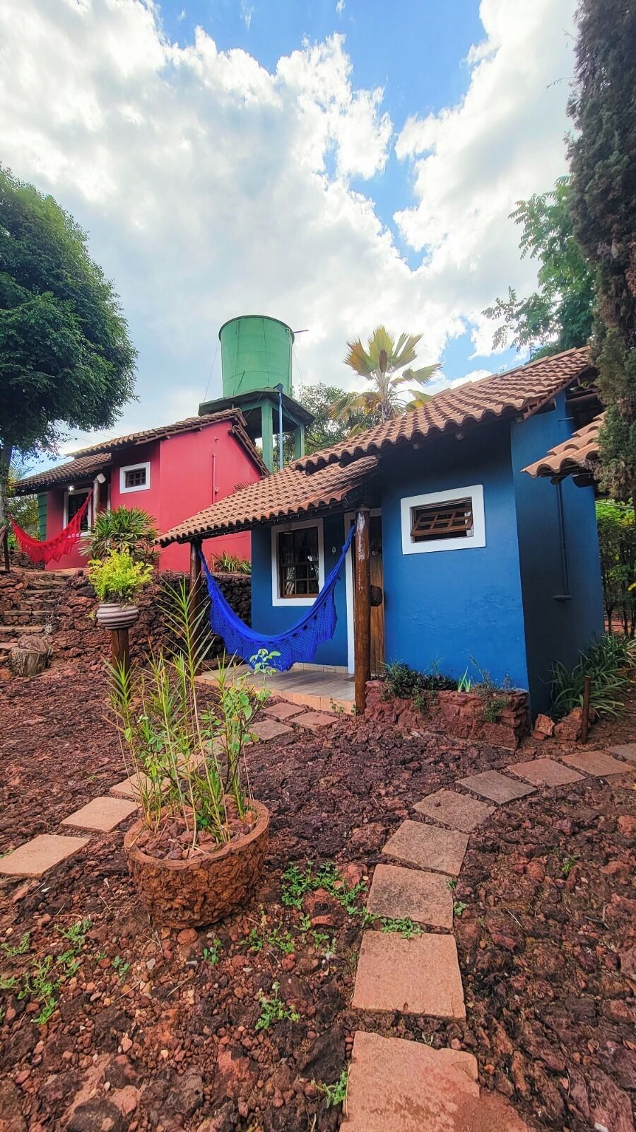 Una casa de campo con fachada roja y azul, un depósito de agua y una hamaca.