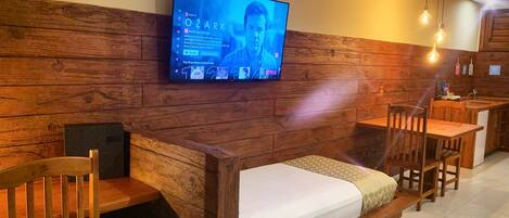 Una habitación acogedora con pared de madera, una cama, un televisor montado en la pared y una pequeña mesa con sillas.