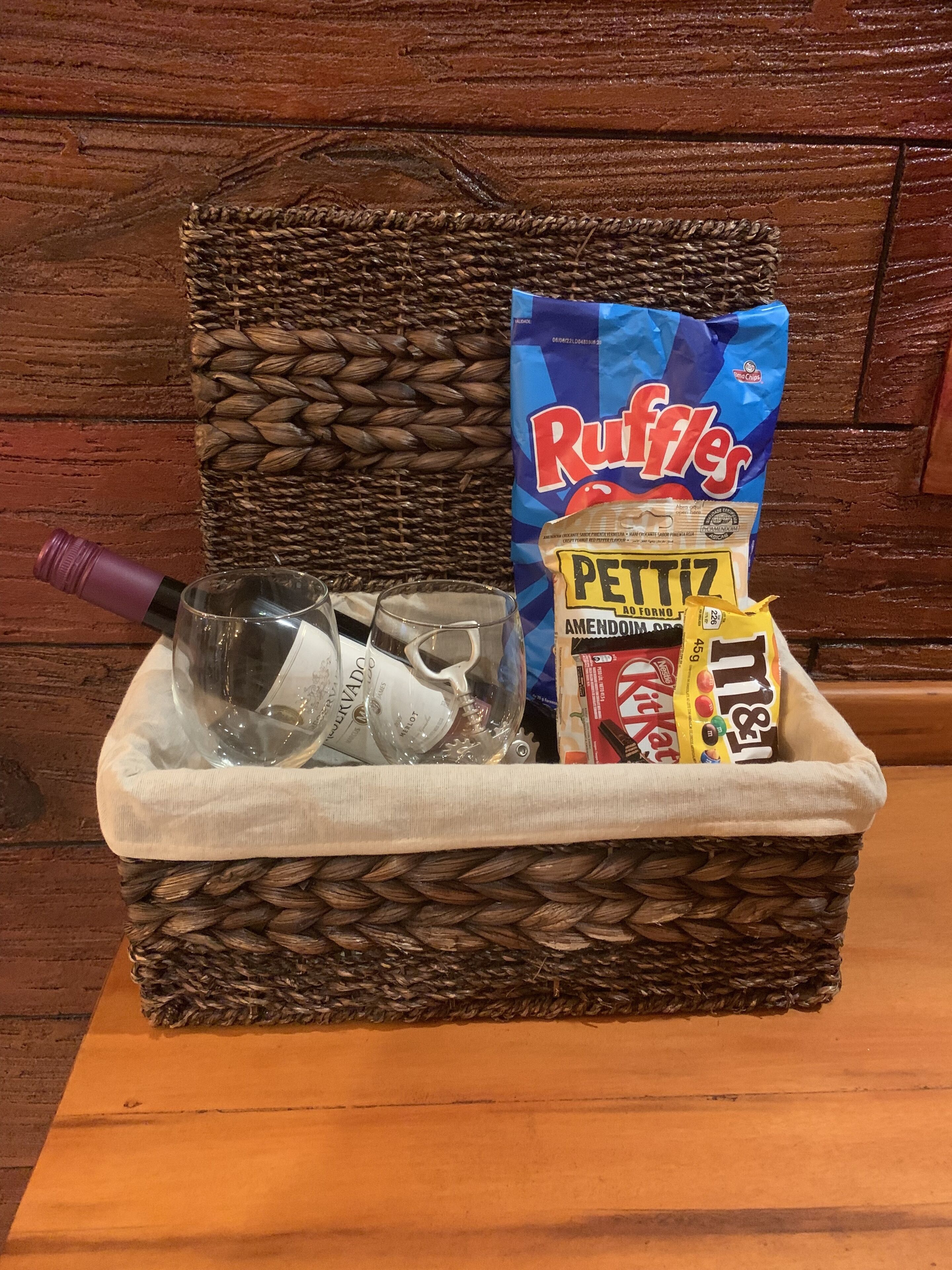 Una cesta de mimbre con una botella de vino, dos copas, una bolsa de Ruffles, una bolsa de Petittz Amendoim y una bolsa de KitKat.