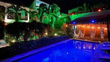 A nighttime view of a resort with a swimming pool, palm trees, and a building with illuminated green and blue lights.