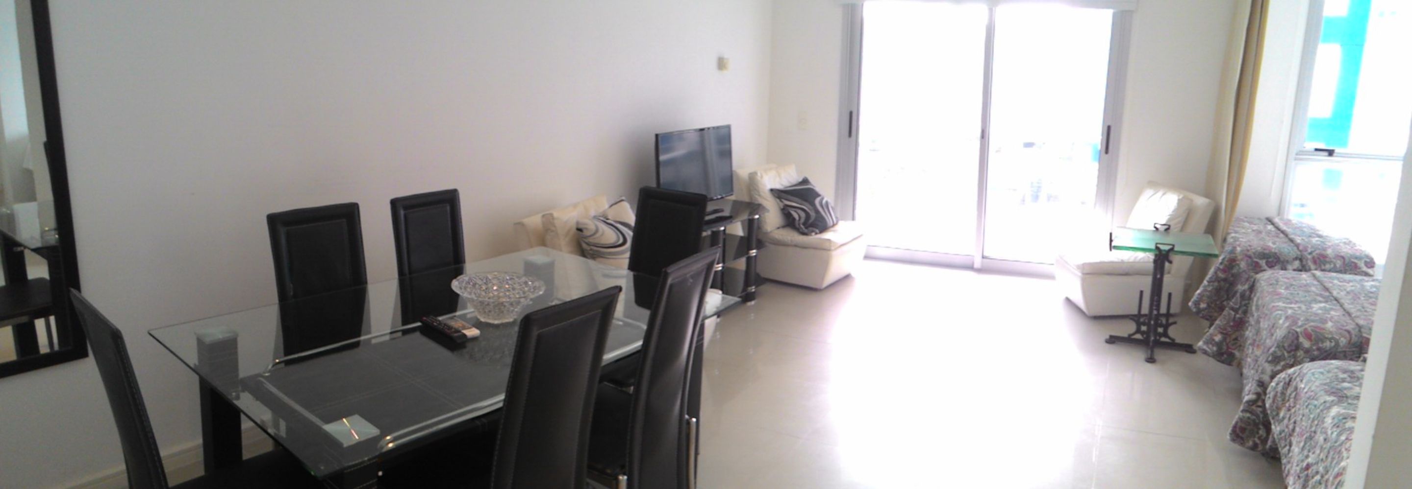 A modern living room with a glass dining table, black chairs, a flat-screen TV, a sofa, and a patterned bed.