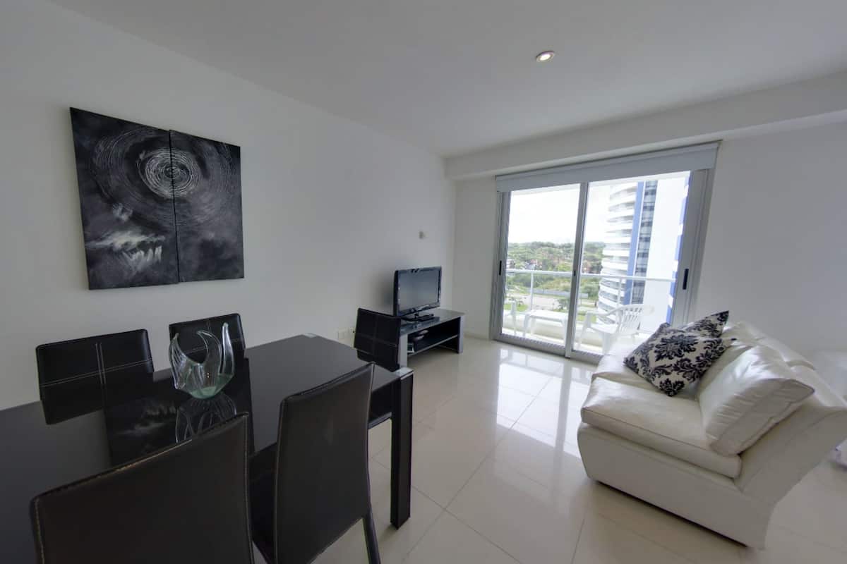 A modern living room with a sofa, a dining table with chairs, a TV, and a large window with a view of buildings.