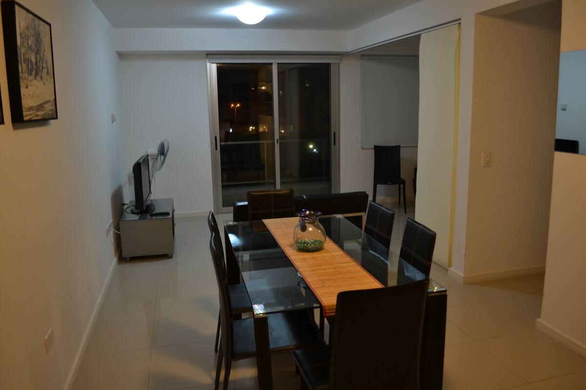 A dining area with a glass table, black chairs, a wooden table runner, and a vase.