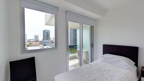 Habitación de hotel con cama, mesita de noche, ventana con vista a la ciudad y puerta corrediza de vidrio que da a un espacio exterior.