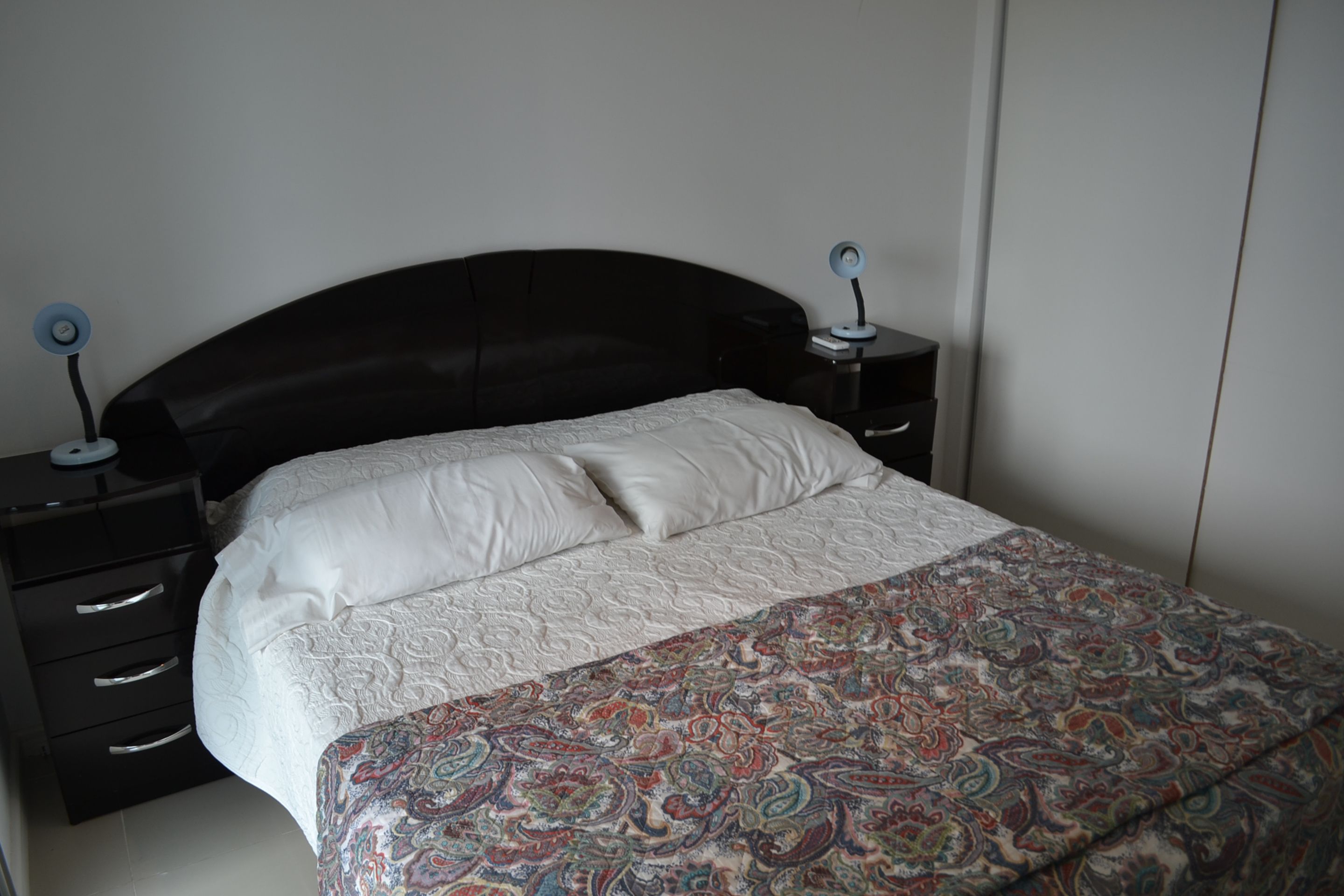 A bedroom with a black headboard, white bedding, and a patterned throw blanket. There are two bedside tables with lamps.