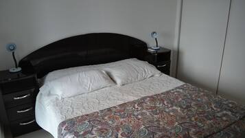 Un dormitorio con cabecera negra, ropa de cama blanca y una manta decorativa estampada. Hay dos mesitas de noche con lámparas.