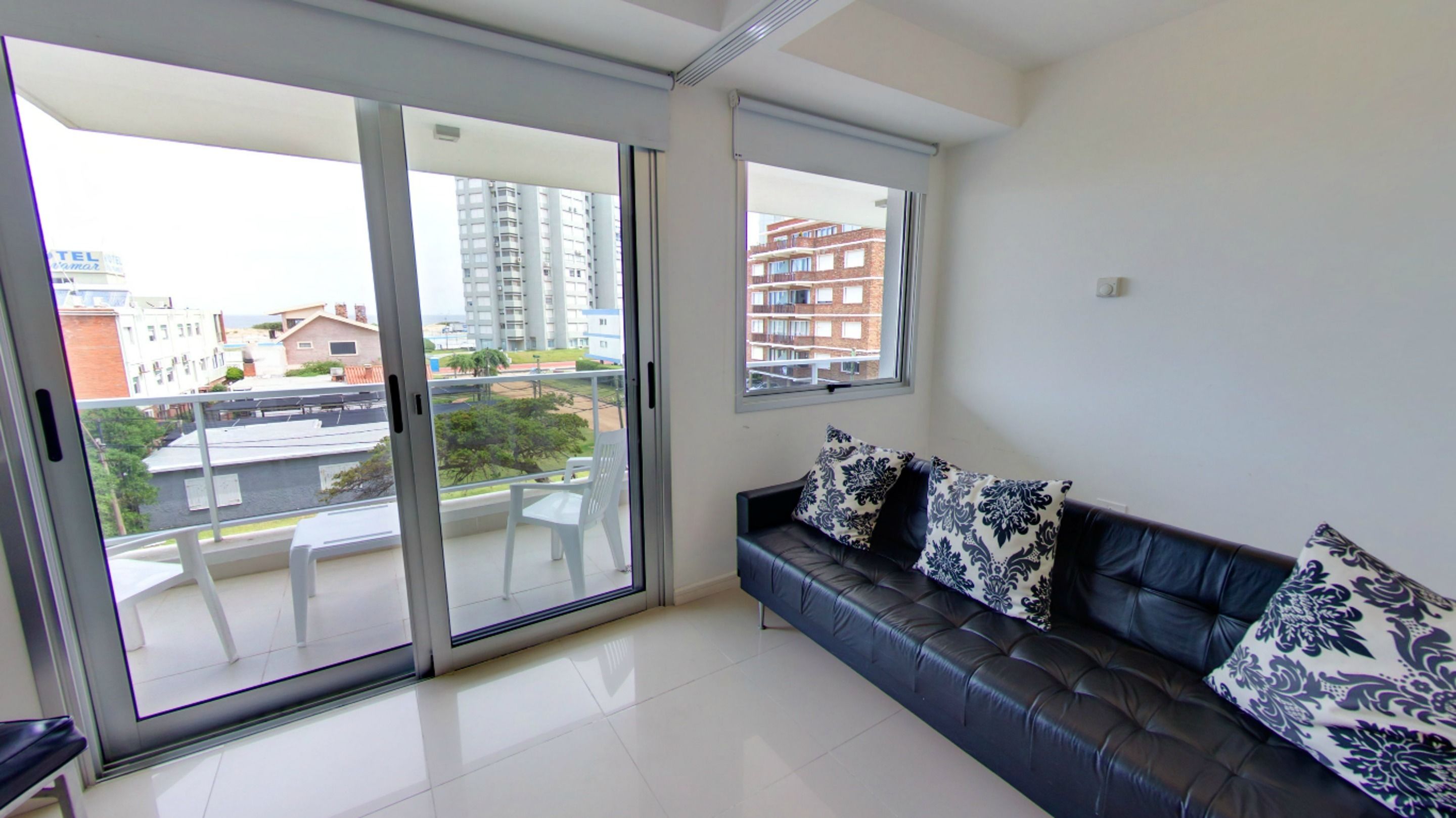 Una sala de estar moderna con un sofá de cuero negro, dos cojines estampados, un ventanal corredero que da a un balcón y vistas de un paisaje urbano con edificios y un río.