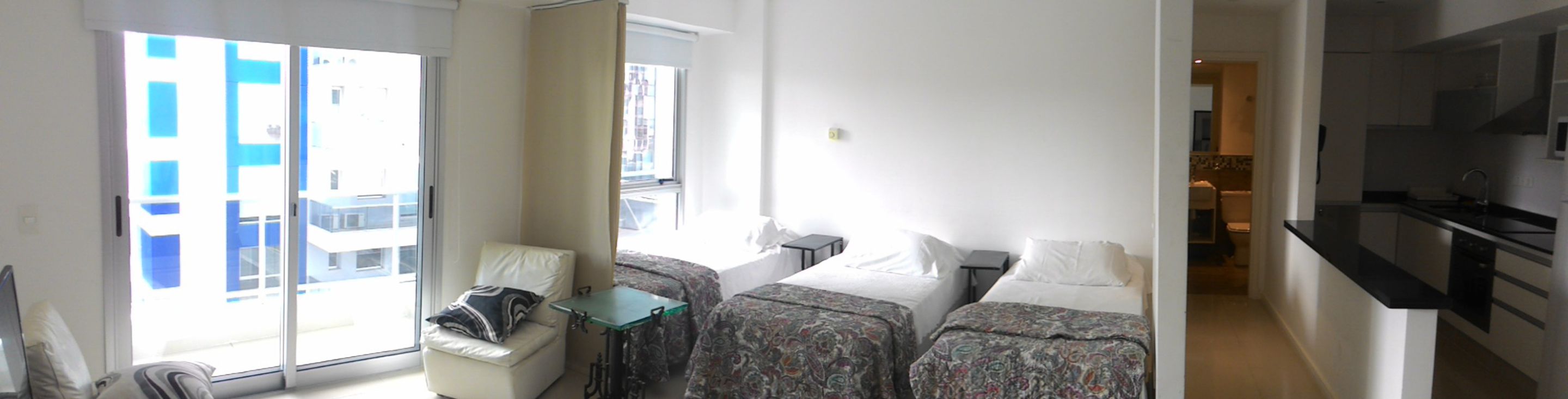 Habitación de hotel con un ventanal grande, una mesita, dos camas con ropa de cama estampada y una kitchenette.