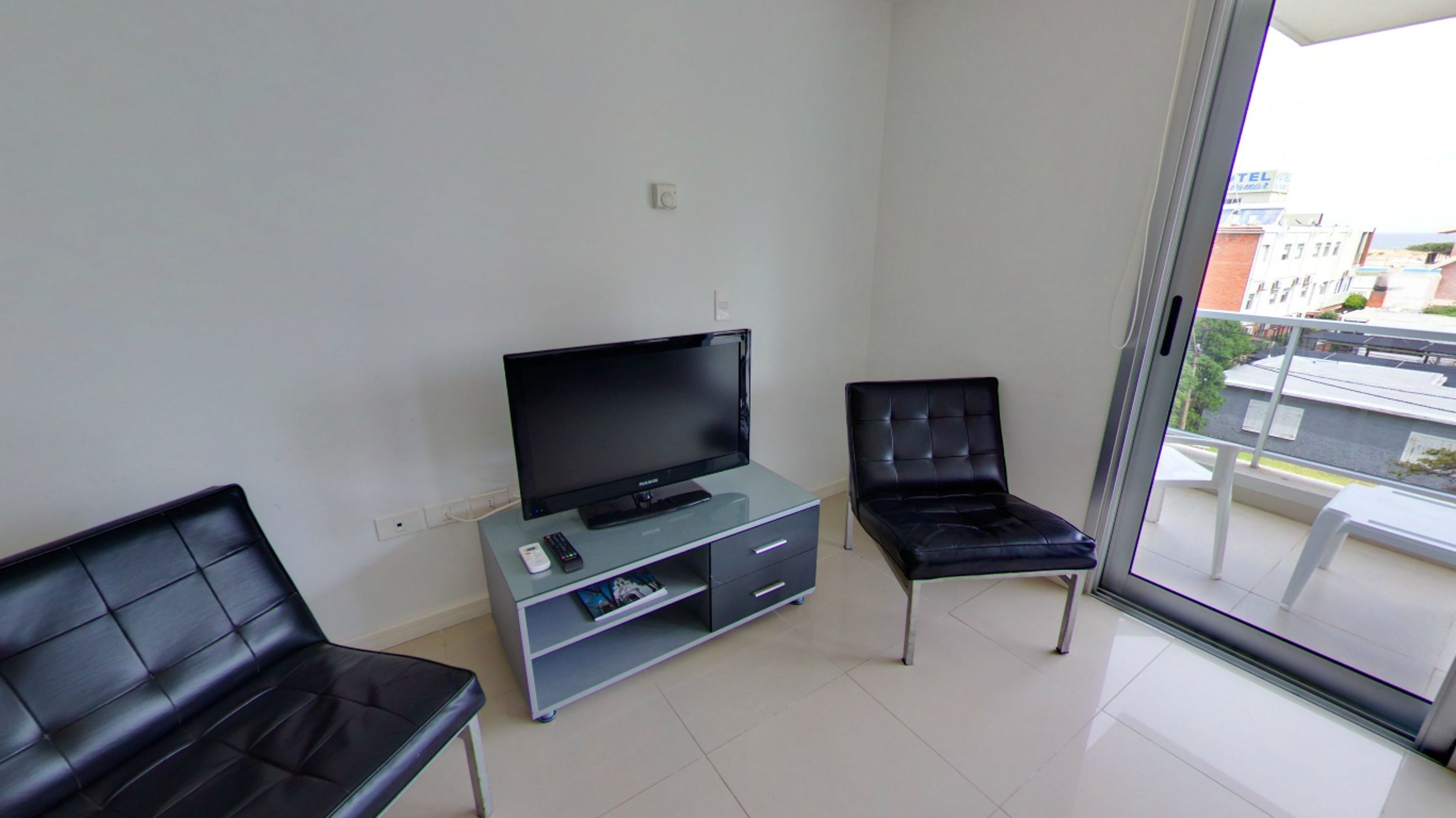 Una sala de estar moderna con una televisión de pantalla plana sobre un mueble de vidrio, dos sillones de cuero negro y un ventanal corredizo que da a un balcón con vistas a la ciudad.