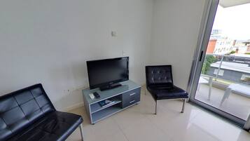 Una sala de estar moderna con una televisión de pantalla plana sobre un mueble de vidrio, dos sillones de cuero negro y un ventanal corredizo que da a un balcón con vistas a la ciudad.