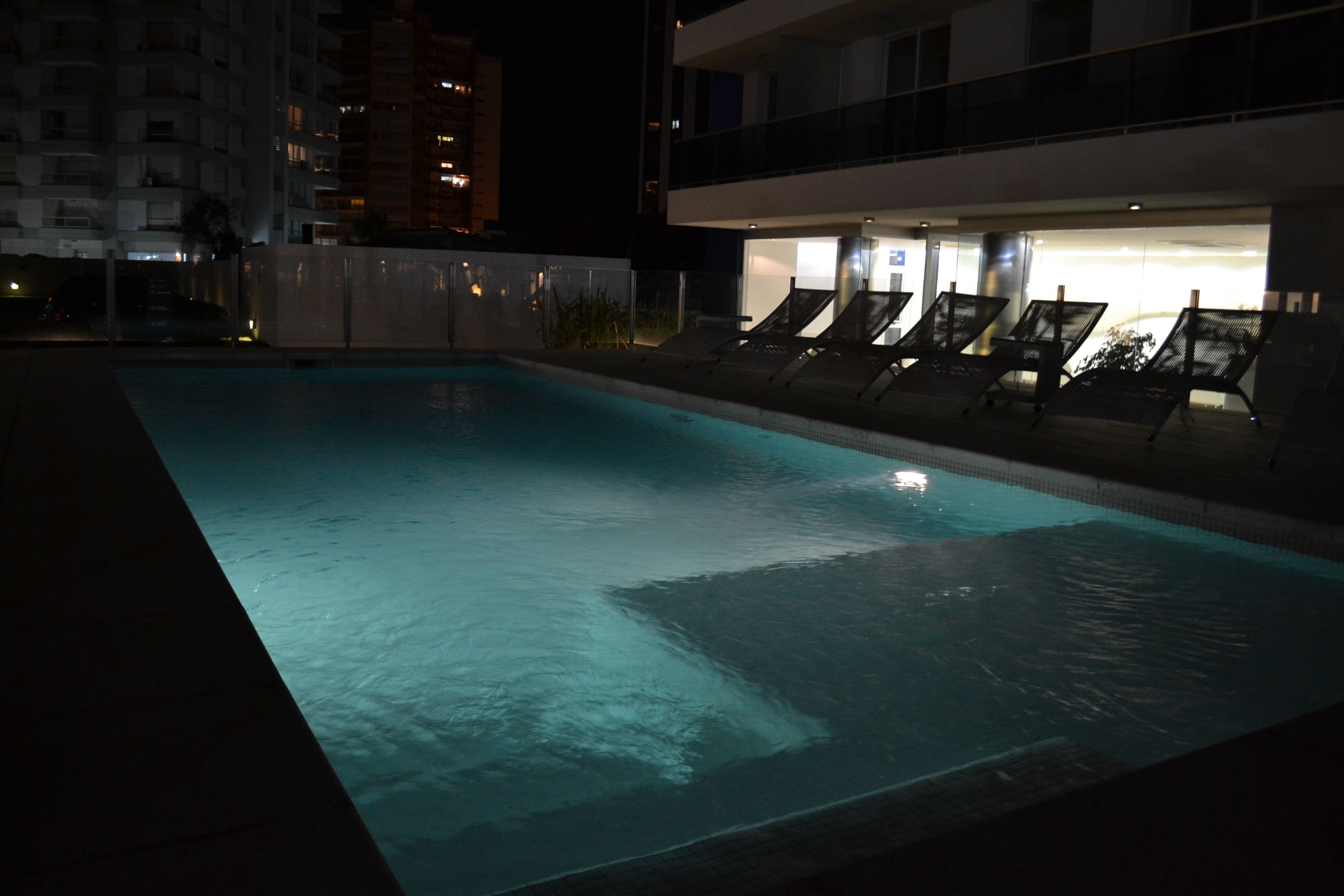 Une piscine éclairée pour la baignade nocturne, dotée de chaises longues, un bâtiment avec des balcons et un ciel nocturne dégagé.