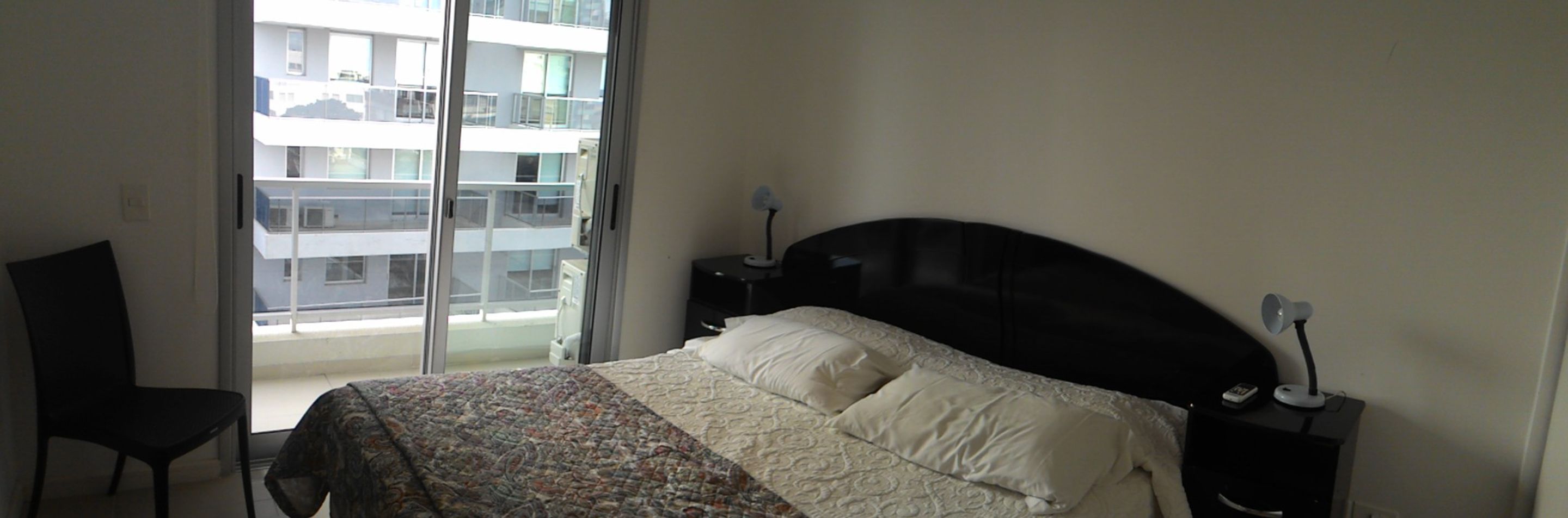 A hotel room with a bed, a chair, a nightstand, and a view of a building through a large window.