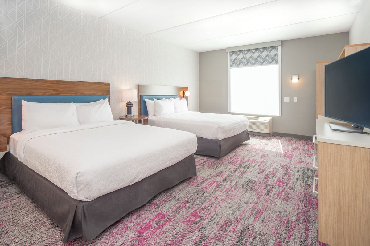 A hotel room with two beds, a large window, a flat-screen TV, and a carpeted floor.