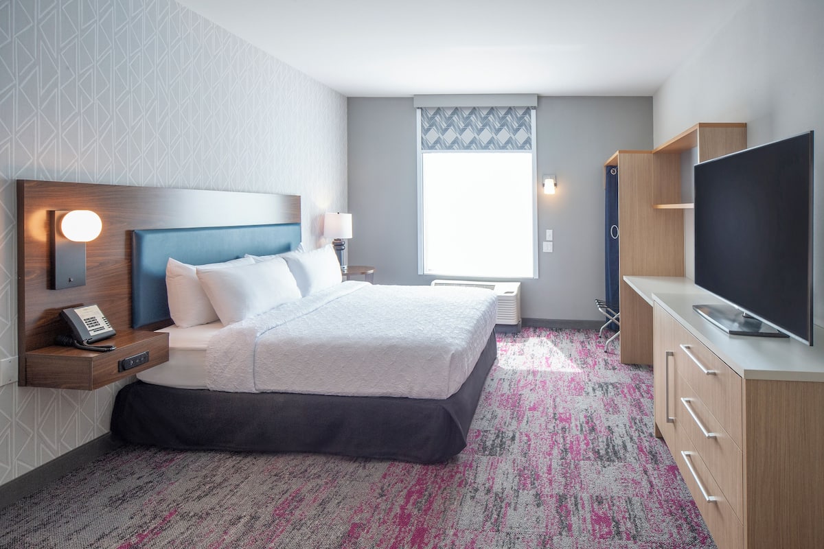 A modern hotel room with a large bed, a bedside table with a phone, a flat-screen TV, and a large window with a view.
