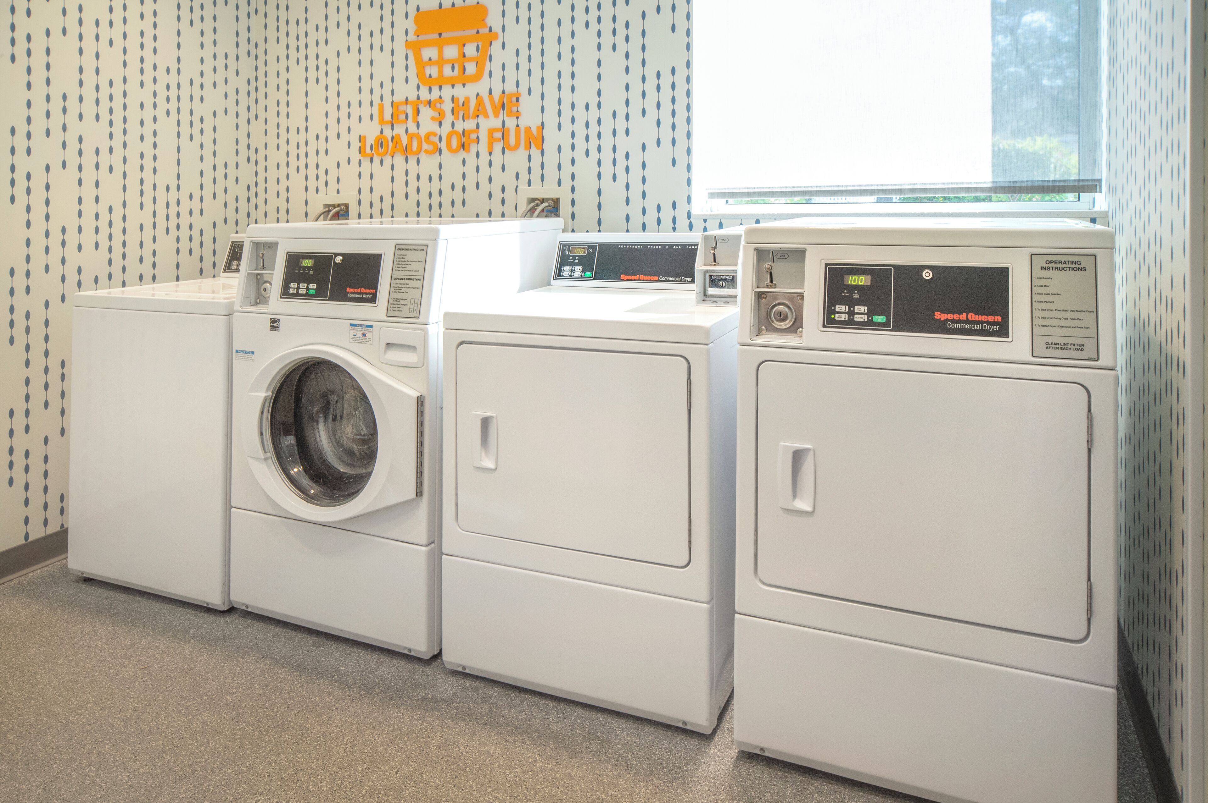A laundry area with three front-loading washers and dryers, a window, and a wall with a motivational quote.